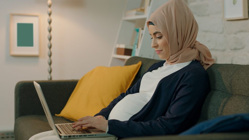 Pregnant woman wearing a headscarf sitting on the sofa working on her laptop