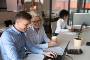 Group of people of various ages, genders and ethnicities working in an office together. Two people are working on the same desk and are smiling, while another colleague is frowning in the background because she may be experiencing symptoms of menopause....