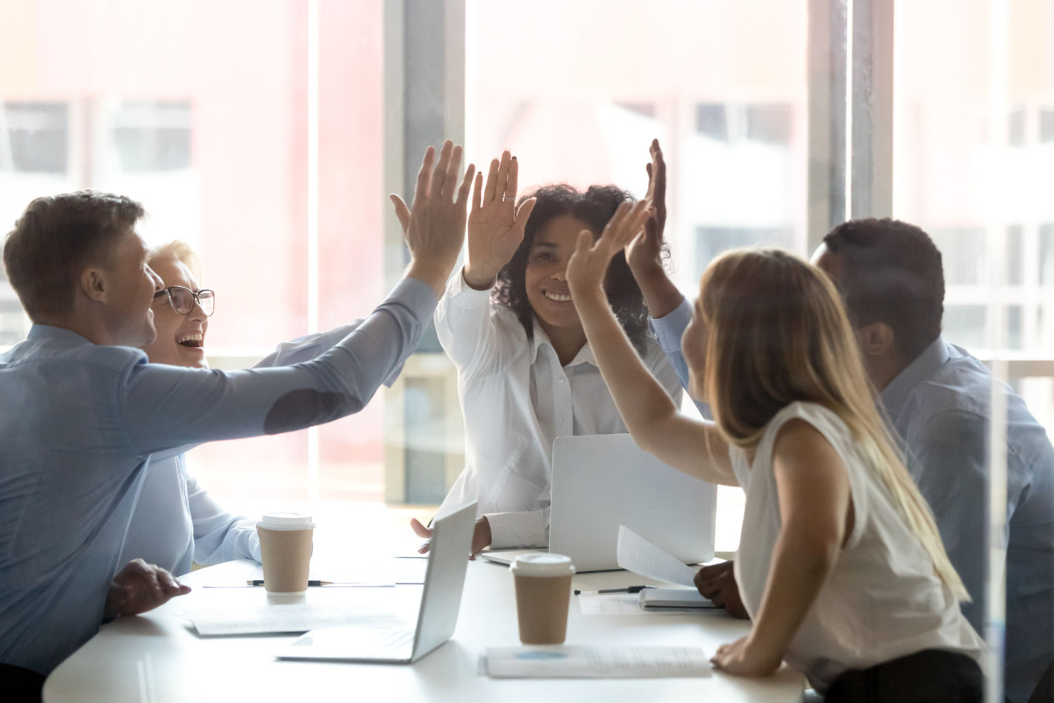Diverse group of office workers high fiving each other, laptops and takeaway coffee cups are on the table in front of them