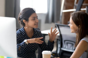 A person talking to another with a laptop screen and coffee, whilst the other person is listening.