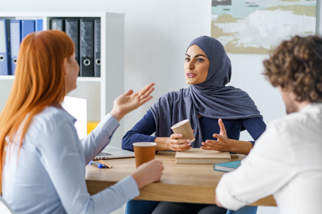 Group of people having a lively conversation over coffee in an office. One of the people is a muslim lady wearing a hijab.