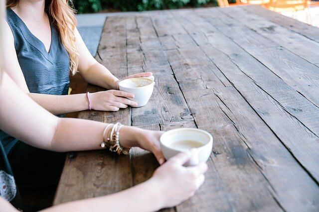 Two people sat at a wooden table with their arms and hands holding coffee cups.