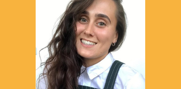Close up of Claudia smiling. They have long brown curly hair, wearing a white shirt with pin stripe dungaree over the top