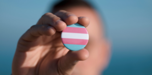 close up of a person's hand, they are holding a button with the colours of the transgender flag in their finger tips