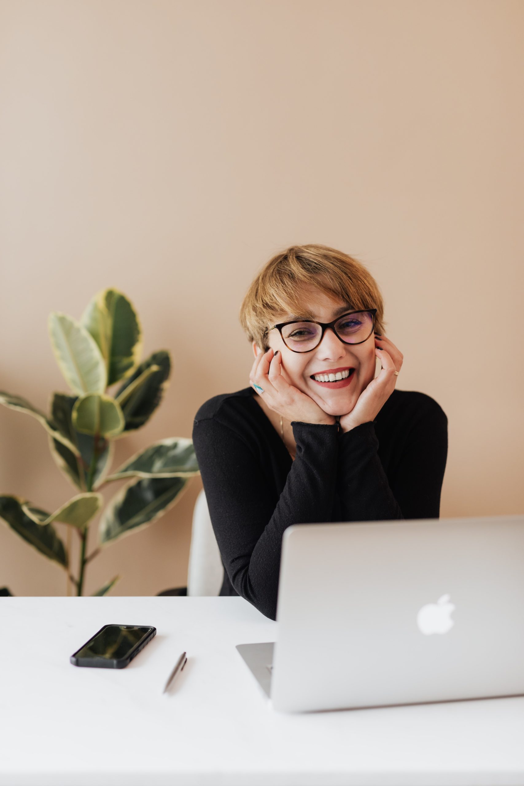 Women working at a laptop, she is smiling at the camera and resting her head in her hands