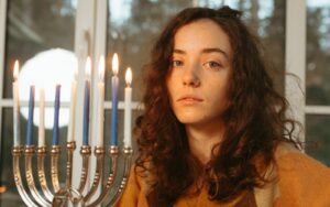 a young jewish women with long brown hair looks into the camera, she is hold a Hanukkah menorah.