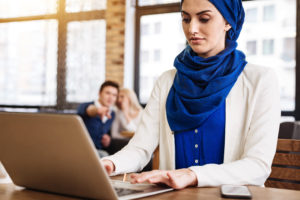 In the foreground a woman is working at a laptop wearing a blue headscarf and top. In the background to colleagues are sitting down, looking like they are talking about her