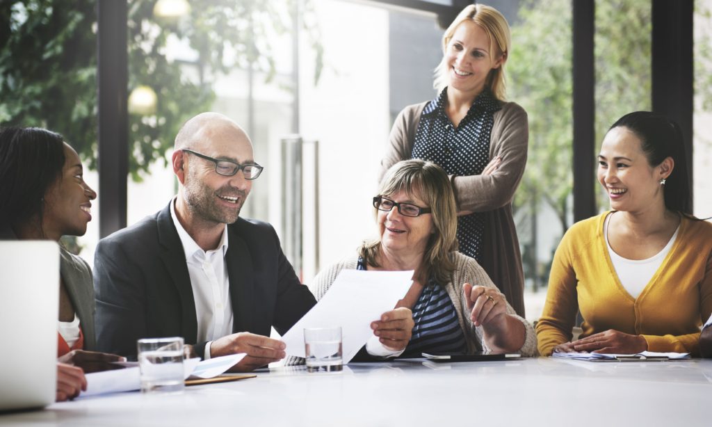 5 colleagues are sitting or standing around a work station. One is reading from a piece of paper, they are listening and smiling