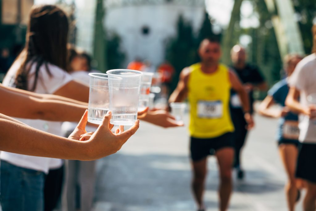 In the foreground you can see people's hands holding out cups of water, in the background runners are about the run past them