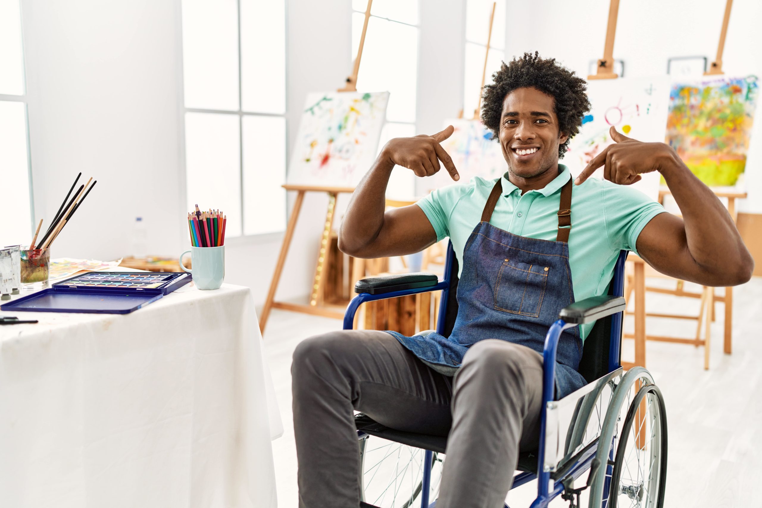 A black person in a wheelchair, they are smiling and pointing both their index fingers at themselves. They are in an artisti studio - their are stands with canvases on in the background