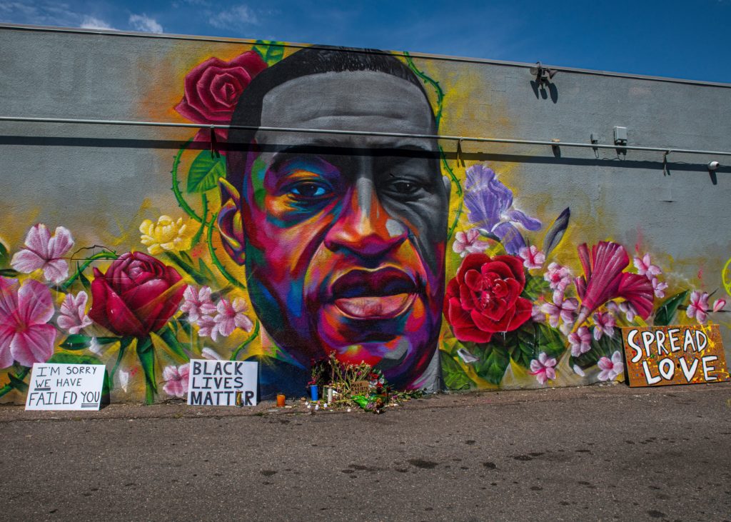 A colourful mural of George Floyd's face with bright flowers around it. There are placards on the ground that read 'Spread Love' and 'Black Lives Matter'