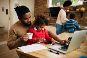 A dad working at his laptop on the kitchen table. His daughter is sitting on this lap. In the background a mother and son are working on a kitchen bench.