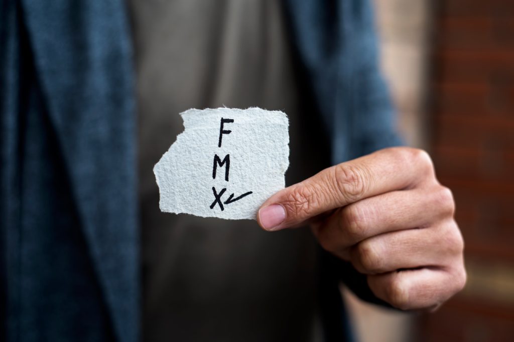 Close up image of a hand holding a piece of paper directly to the camera - the paper has the letters F, M and X, written vertically down the page. There is a tick next to the X