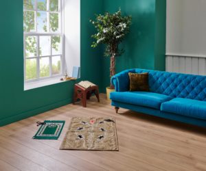A room with a green wall and a large window, there are prayer mats on the wooden floor and a bright blue sofa behind them.