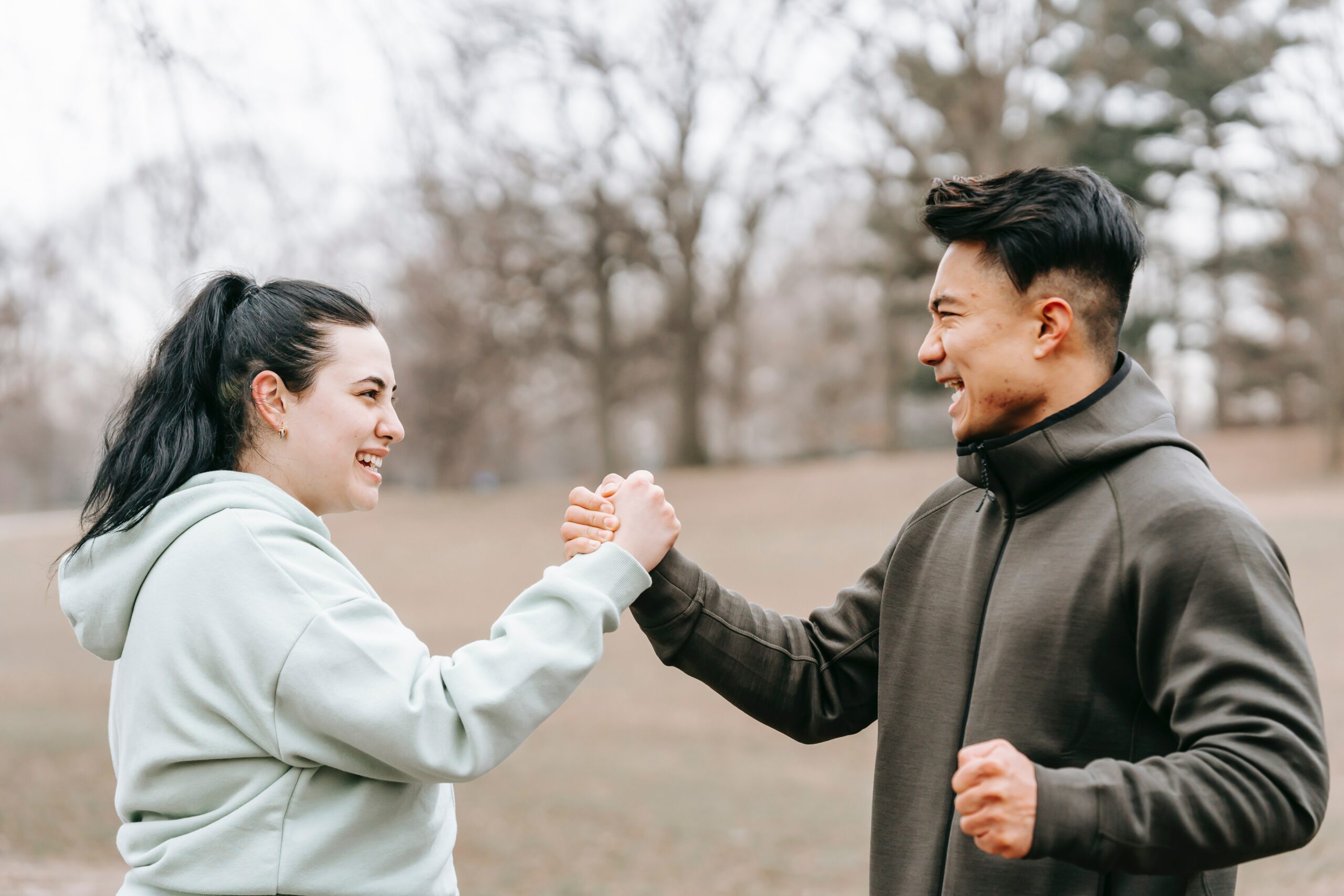 An athlete clasping the hand of their colleague, both are wearing sporty clothes, smiling to one another and standing in a park. They seem happy and excited to be working together.