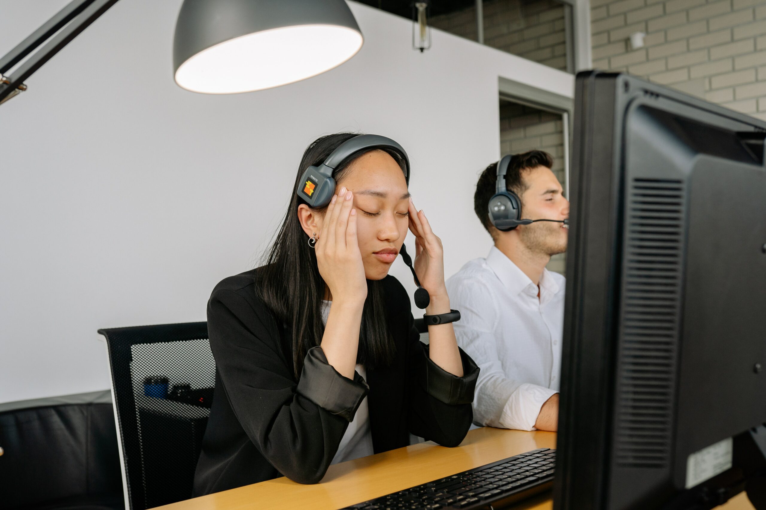 A person sitting at a desk with a screen and headset, with eyes closed, rubbing their temples