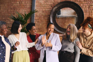 Diverse group of women celebrating international women's day. They are all in a fancy brickwork office. One woman is happily clapping her hands and all the woman are joining in on the laughter.