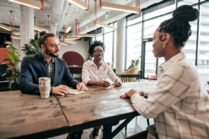 Three office workers are sitting in an open plan office with a big window. There are plants everywhere. Two older colleagues are sat at a long wooden table, mentoring a younger employee.