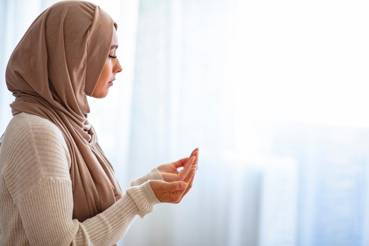 A Muslim woman wearing the headscarf is praying in her head office's prayer room. She is raising her hands in Dua, which means she's invoking Allah. Her hands are turned upwards and cupping the air.