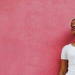 Black female wearing her hair in a headscarf leans against a bright pink wall.
