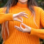 A person with long hair and wearing an orange jumper doing sign language
