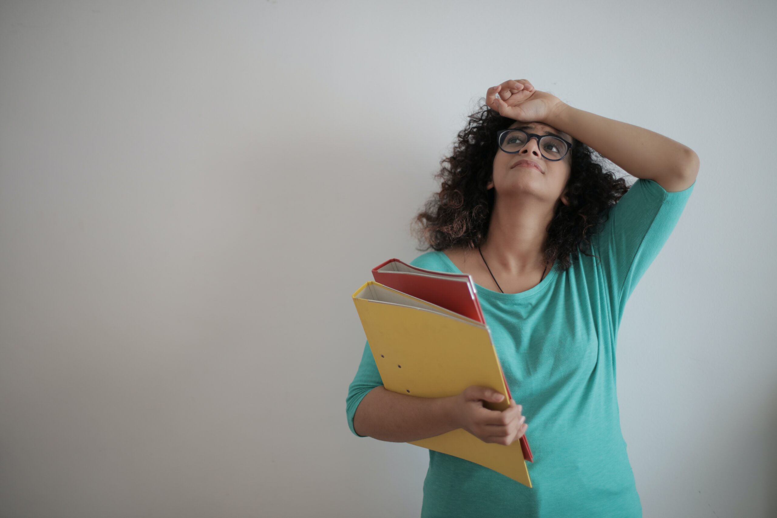 A feminine presenting person wearing a turquoise top, standing against a white wall holding some files with one hand raised to their head in exasperation