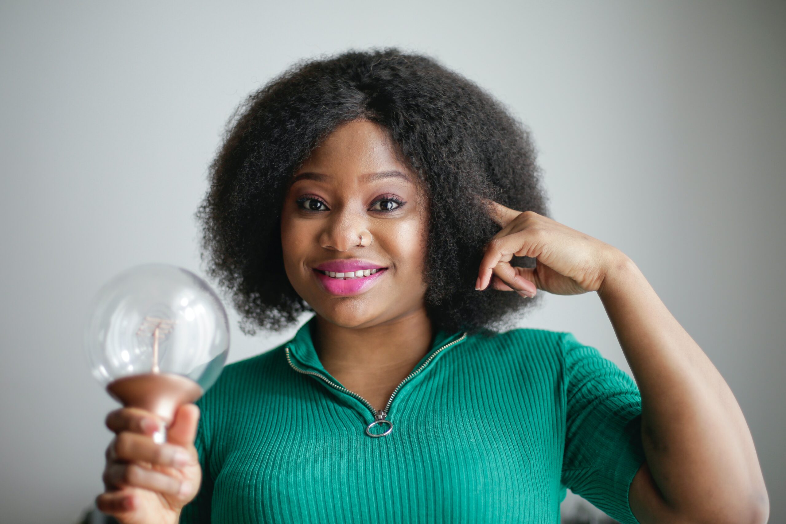 Black, feminine presenting person with a green polo shirt, holding a light bulb and pointing to their head