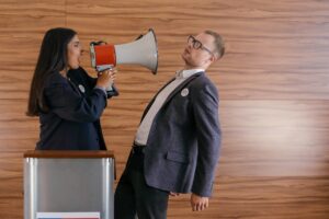 A feminine presenting person of colour with a megaphone directed towards a White masculine presenting person shouting something, both in work suits in an office environment