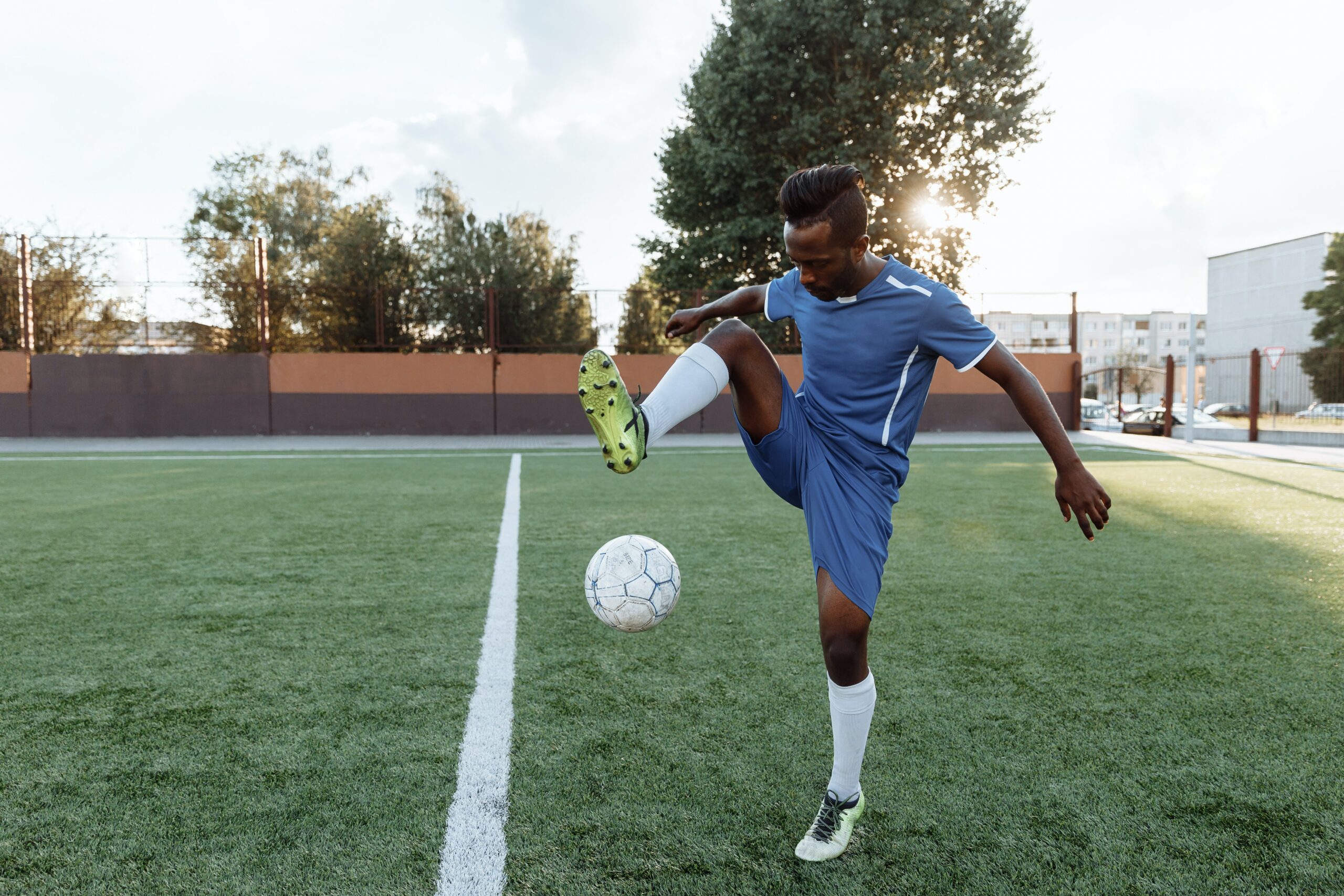 A masculine presenting person of colour in a football kit on a green grass pitch playing with a football