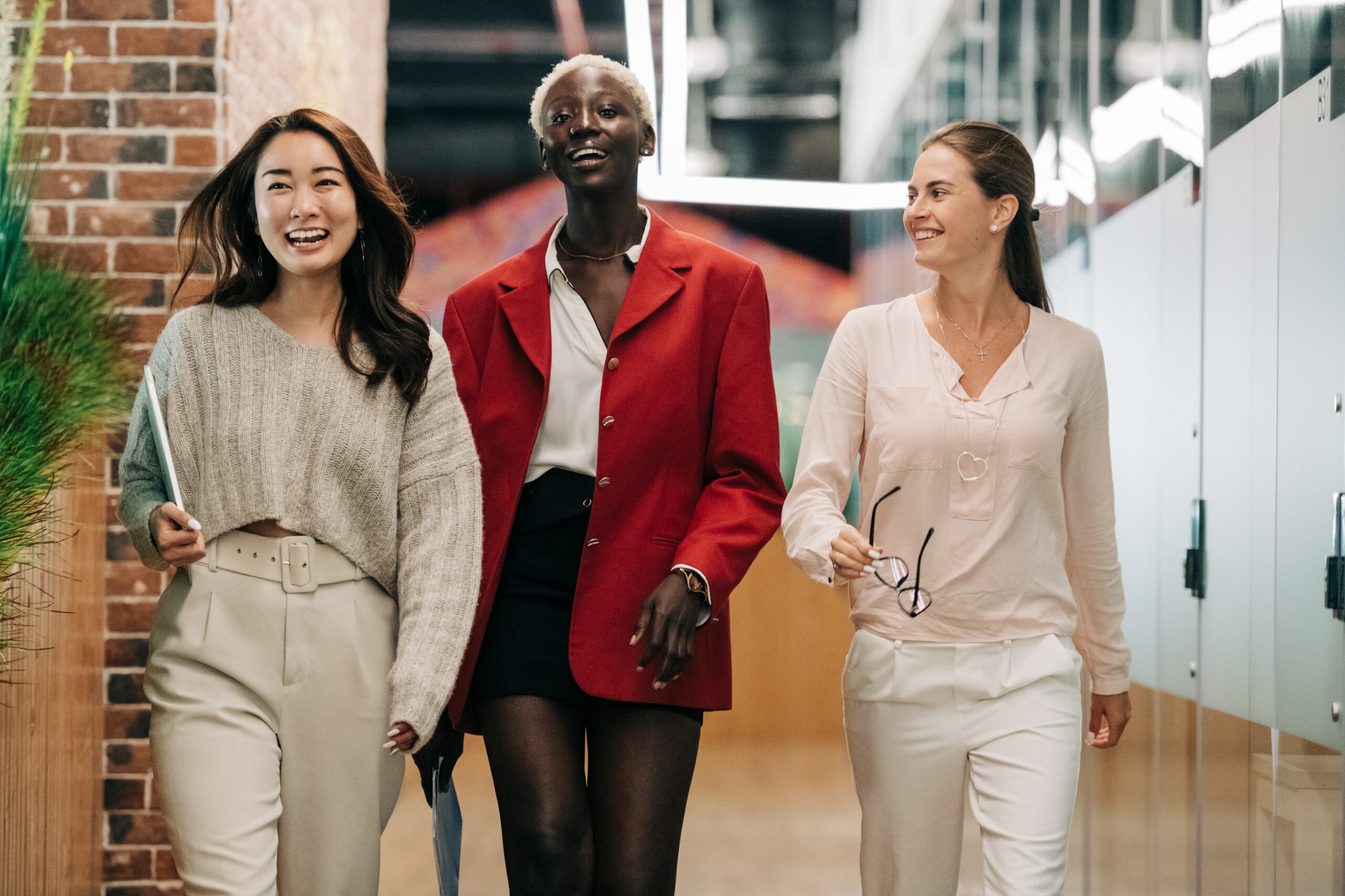 Three feminine presenting people walking down an office corridor in work attire.