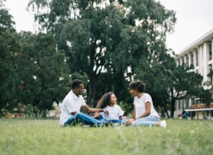 A black family of two adults and a child sitting on the grass in a park