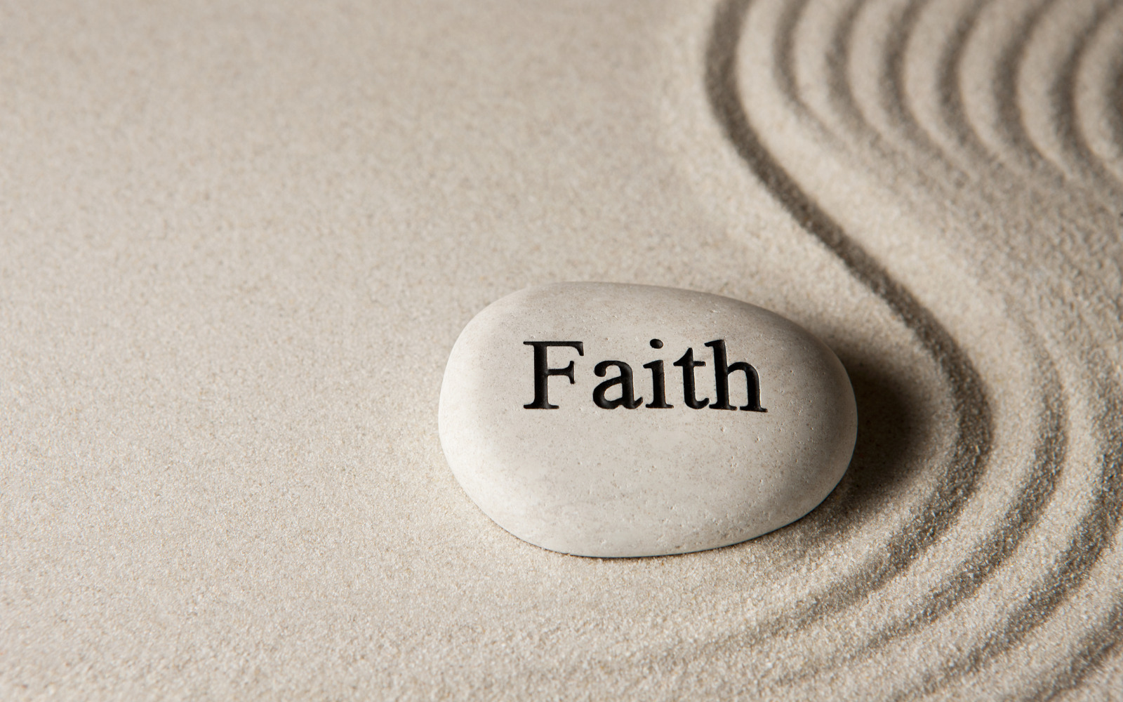 A pebble on carefully sculped sand. The pebble has the word 'Faith' written on it.