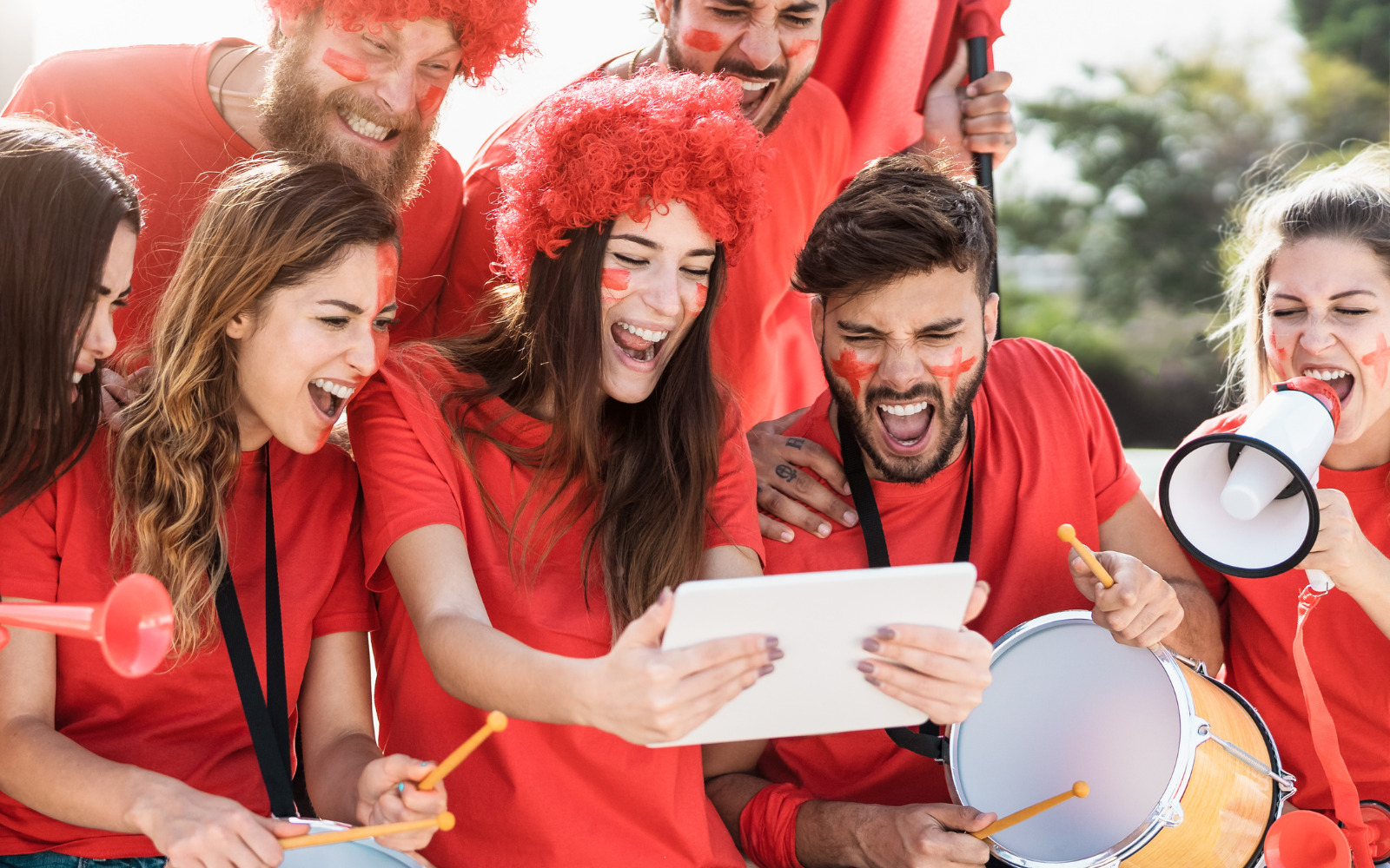 A group of sport fans in red cheering at a handheld tablet one of them is holding.