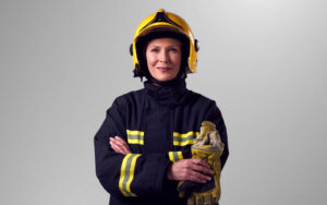 Feminine presenting fire service worker in navy suit with yellow stripes, they are wearing a yellow helmet and are folding their arms