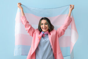 Trans person holding up the Trans Pride flag and smiling