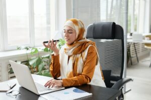 Muslim person wearing a hijab sitting at a desk working at a laptop, while speaking on the phone