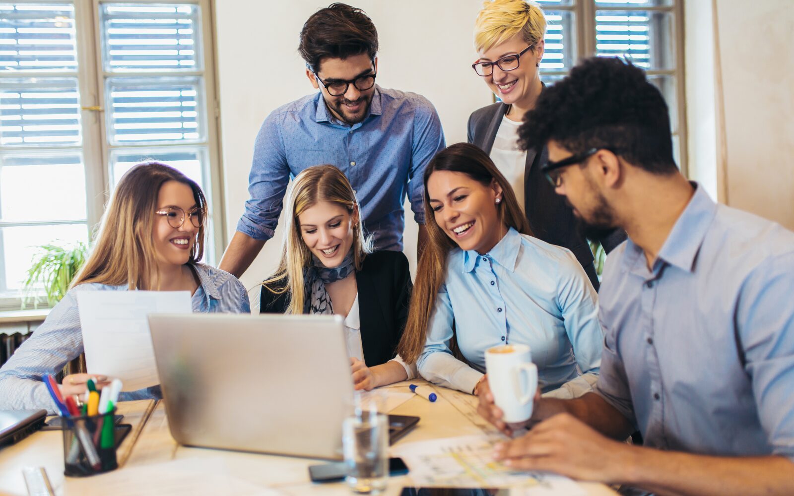 A group of diverse colleagues sitting around a laptop on table and smiling together