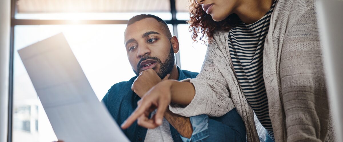 Two people reviewing their current policies. A masculine Black person is holding up a white piece of paper for the other person to read.