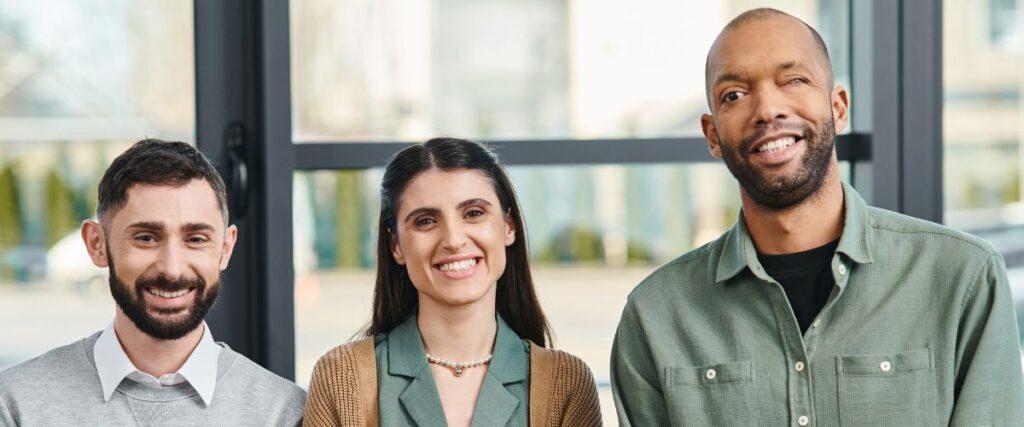 A diverse group of three colleagues standing together and smiling in an office setting with large windows in the background. The individual on the left has a beard and wears a light gray sweater over a white collared shirt. The person in the center has long dark hair and wears a green blazer with a pearl necklace. The individual on the right has short hair, wears a green shirt, and has a visible facial difference. The group exudes a friendly and inclusive atmosphere.
