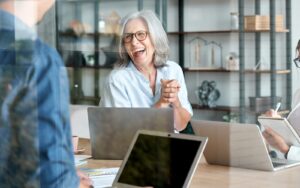 Person in glasses laughing in a meeting, there are several laptops on the desk in front of them