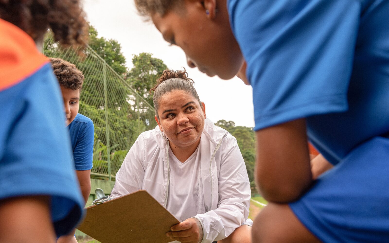 A coach huddled in a circle with their students. They are holding a clipboard and smiling at their students.
