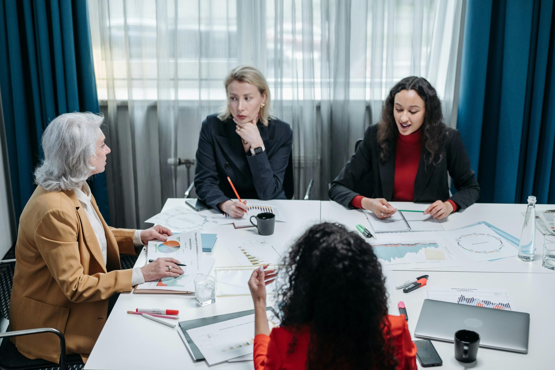 Four women sitting around a table that is covered in paper. They are wearing business outfits and formal attire and are in deep conversation.