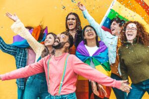 Image shows a diverse group of people in bright clothing with a Pride flag and confetti